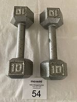 Photo of two 10-pound hex dumbbells with metal finish and visible wear, placed on a white surface with auction lot number.