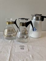 Photo of two Pyrex carafes with black handles, one with horizontal gold stripes and one with gold starburst pattern, plus white Corningware coffee pot with blue floral decoration and metal collar lid.
