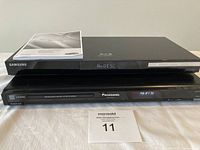 Two DVD players stacked: Samsung Blu-ray player on top, Panasonic DVD player below, with Samsung manual on top.