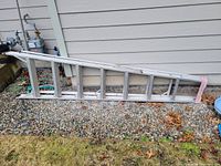 Full view of the aluminum step ladder laying on gravel beside a house exterior wall. Shows full length and construction details of the ladder.