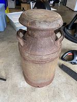 Full front view of antique metal milk can on concrete floor, showing overall shape, handles, and surface rust.