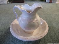 Porcelain pitcher with embossed floral and swirl decorations sitting inside matching bowl on carpeted floor.