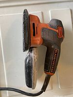 Side view of Black & Decker handheld electric sander showing orange and black body with power cord.