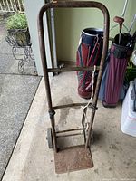 Full view of rusted vintage dolly showing metal frame, handle, and base plate with two wheels, placed on concrete floor near golf bags.