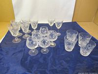 Full lot of 15 crystal glasses including tumblers, stemmed wine glasses, and brandy glasses displayed on a blue cloth.