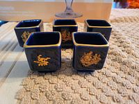 Five dark blue square miniature Limoges cups arranged on textured surface, each with gold classical figure decoration