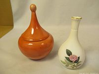 Two vases against plain background, one orange ceramic with pointed lid, one white floral Wedgwood vase