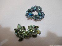 Two vintage glass rhinestone brooches on a white background showing front with blue and green stones, circular and branch shapes.