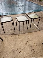 Three square outdoor tables with glass tops and metal frames standing side by side on textured concrete near a covered pool.