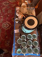 Photo showing a teal Dash air fryer, multiple teal coffee mugs arranged, pitchers including white ceramic and clear glass, and smaller kitchen decor pieces on a table with a patterned rug beneath.