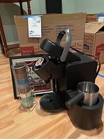 Kerrigan coffee maker with frothing pitcher, metal stand with four stacked espresso cups, and framed coffee print on floor next to cardboard boxes.