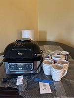 Ninja Foodi 5-in-1 Indoor Grill on a table with 13 assorted ceramic coffee mugs behind it.