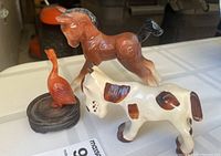 Three ceramic animal figurines on a surface: a brown horse, a white dog with brown spots, and an orange bird on a small black wooden base.