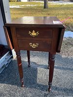 Full view of wooden end table showing two drop-leaf sides, four turned legs, and two drawers with brass handles.