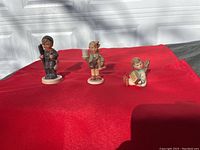 Three Hummel figurines placed on a red cloth in natural lighting.