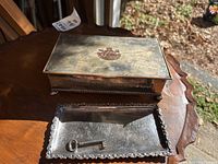 Photo showing the rectangular metal box with crest on lid, the rectangular metal tray with scalloped edges, and the small antique key on the tray. Set on a wooden surface with natural light.