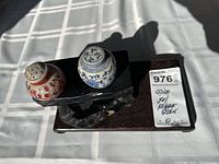 Photo showing two ceramic shakers with floral designs on a wooden and black stone display stand under natural light.