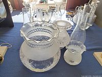 Photo showing all glass items including frosted crystal globes with floral etching and clear glass chimneys on a blue cloth surface with sunlight reflecting.