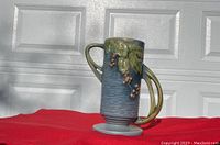 Roseville pottery vase on red cloth, side angle showing large curved handle and leaf and berry raised decoration