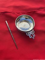 Two sterling silver items on red fabric background; an ornate pointer and a small trinket dish with pierced handle.