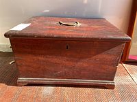 Front view of the wooden storage chest showing brass handle, keyhole and molded base trim.