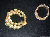 Two vintage bracelets on black background, one gold-tone double strand with translucent beads, one gold-tone multi-row rhinestone bracelet with stretch band.