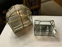 Two glass display cases on white surface, one gold framed with curved top and one silver framed rectangular case.
