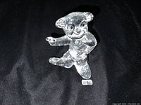 Clear glass clown figurine standing on one leg with one arm pointing out, placed on a black fabric background with BODA label nearby.