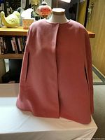 Front angled view of dusty rose/pink cape on mannequin against indoor background with lamps and decorations.
