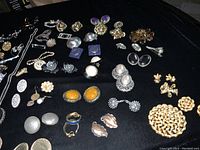 Top down view of assorted jewelry items laid out on black cloth showing earrings, brooches, pins, and necklaces in different metal tones and designs.