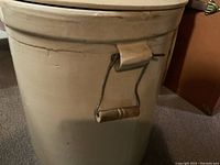Front side view of off-white ceramic crock with a metal handle featuring a wooden grip, no visible cracks but minor wear.