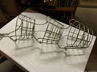 Three silver metal wireframe heavy duty hanging baskets connected by chains shown on white surface