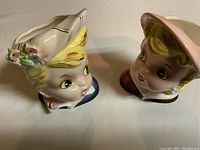 Front view of two porcelain head vases showing facial features and hats with floral accents.