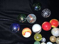 Close up of 11 large marbles with various colors including red, black with white spots, transparent, blue, orange swirl, and white.