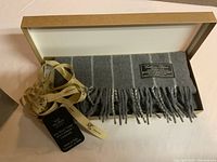 Photo of a grey men's scarf neatly folded inside a beige box with a ribbon and tags indicating it is a cashmere blend by Brooks Brothers.