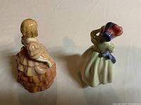 Back view of two Royal Doulton figurines: Rose in peach dress and Babie in pale green dress adjusting bonnet.