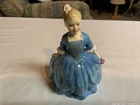 Front view of the porcelain figurine depicting a young girl in a blue dress with a white flower in her hair, sitting position.