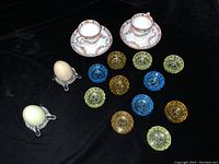 Photo of all items including two mini cups and saucers, eleven colored glass salt dishes in blue, amber, and green, and two clear glass egg holders with eggs.