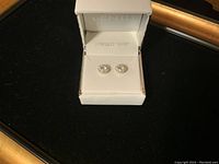 Pair of sterling silver earrings inside a white box with padded interior labeled 'Sterling Silver'. Earrings round with clear stones in center and halo.