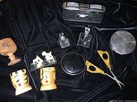 Top view of vintage items arranged on black cloth showing opera glasses, egg cups, compacts, scissors, miniature perfume bottles, dog figurines, and wooden cup/stamp.