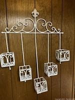 Front view of the white metal wall planter with decorative scrollwork and four hanging square holders attached to a wood background.
