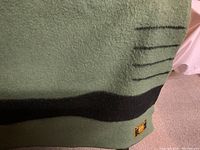 Close-up of blanket showing dark green wool fabric with a wide black horizontal stripe and a sewn-on label patch with a logo.