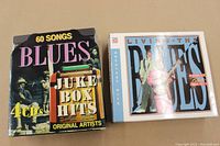 Photo shows two boxed CD sets: on left, the 4-CD set "Blues Juke Box Hits" featuring 60 songs by original artists. On right, the 3-CD set "Living the Blues" featuring 36 tracks by various classic blues musicians.