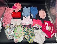 Photo showing all 14 baby bathing suits arranged on a black surface, highlighting variety in colors, styles, and patterns including ruffles and unicorn designs.