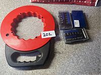 Red circular fish tape with ergonomic handle alongside two compact blue ratchet socket and bit sets laid out on carpet.