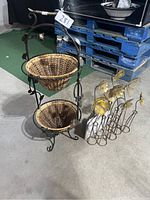 Two-tier wicker basket stand with decorative metal frame and a set of metal floral garden stakes placed on concrete floor near blue pallets.