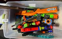 Top-down photo showing multiple colorful toy dart guns and toy rifles piled inside a plastic storage bin.