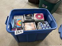 A large blue plastic bin containing many assorted music CDs in jewel cases with visible album cover art.