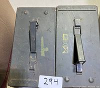 Top view of two large military ammunition cans with black metal handles. The left can features a yellow gun stencil, the right can has another stencil with cartridge information.