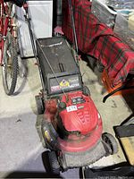 Red Troy-Bilt lawn mower with rear grass collection bag, 21 inch deck, placed beside bike and other household items